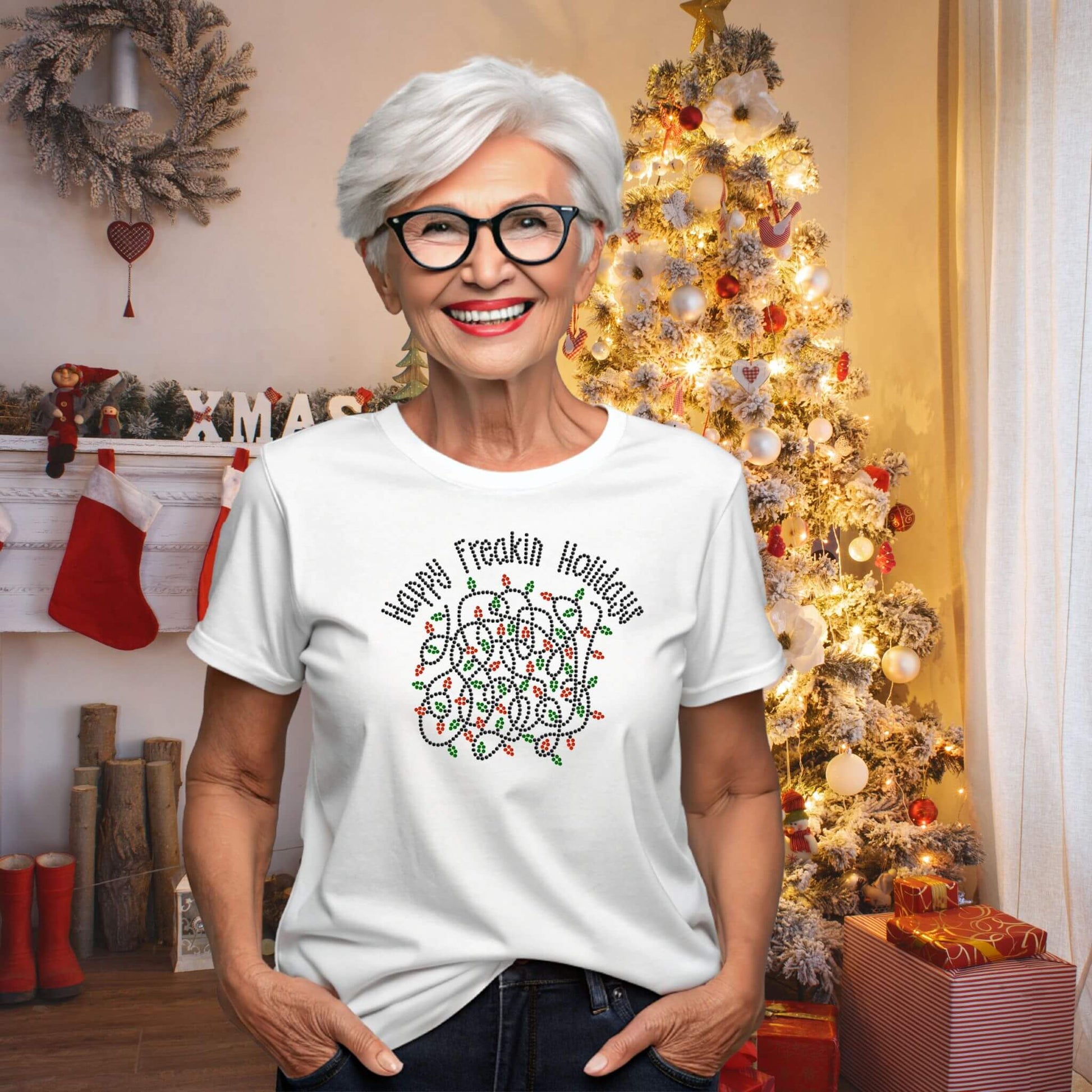 Smiling woman wearing a white “Happy Freakin Holidays” T-shirt with a rhinestone-style tangled Christmas lights design, standing in front of a decorated holiday tree and cozy fireplace.