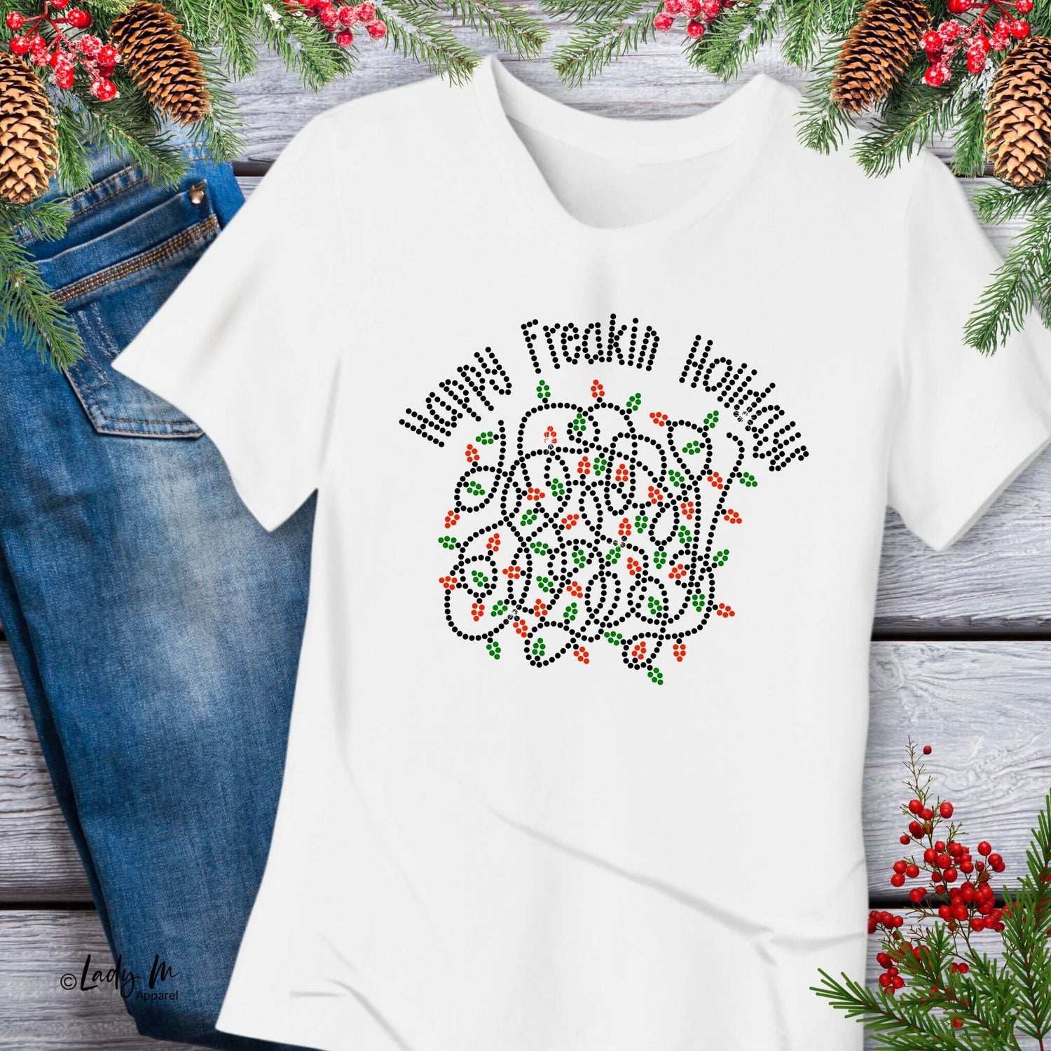 White “Happy Freakin Holidays” T-shirt with a rhinestone-style tangled Christmas lights graphic, displayed flat with denim jeans and surrounded by pine branches, berries, and pinecones on a rustic wood background.