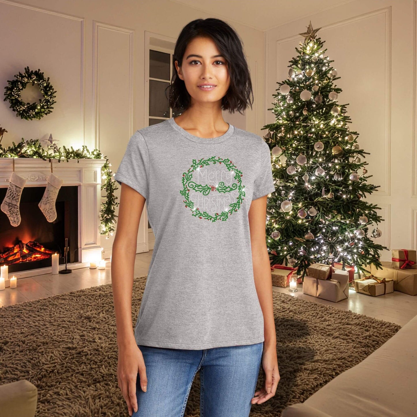 Woman wearing a gray T-shirt with a rhinestone-style Christmas wreath and holly design, standing in a cozy holiday living room with a lit tree and fireplace.