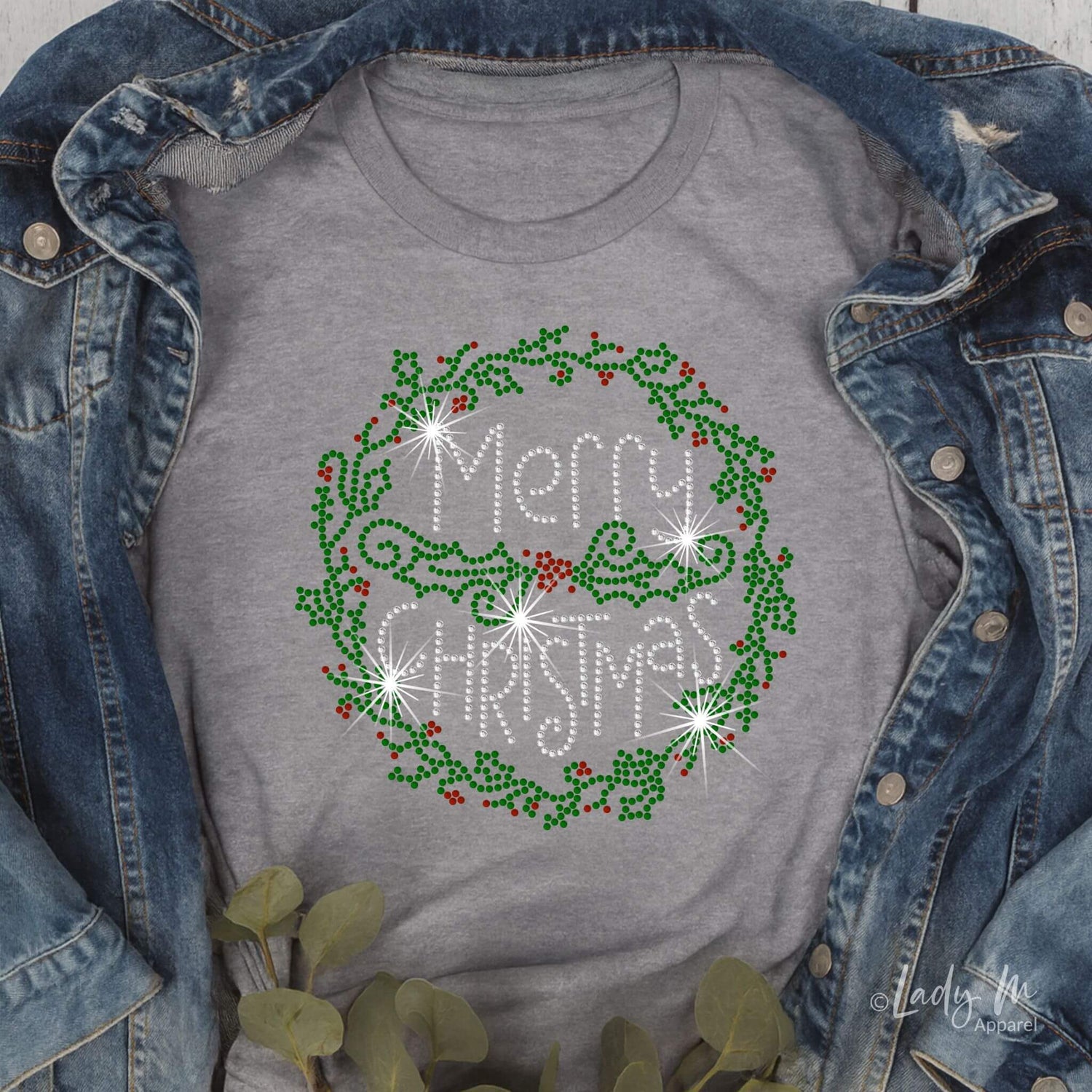 Flat lay of a gray T-shirt with a rhinestone-style “Merry Christmas” holly wreath design, styled with a denim jacket and greenery on a rustic wood background.